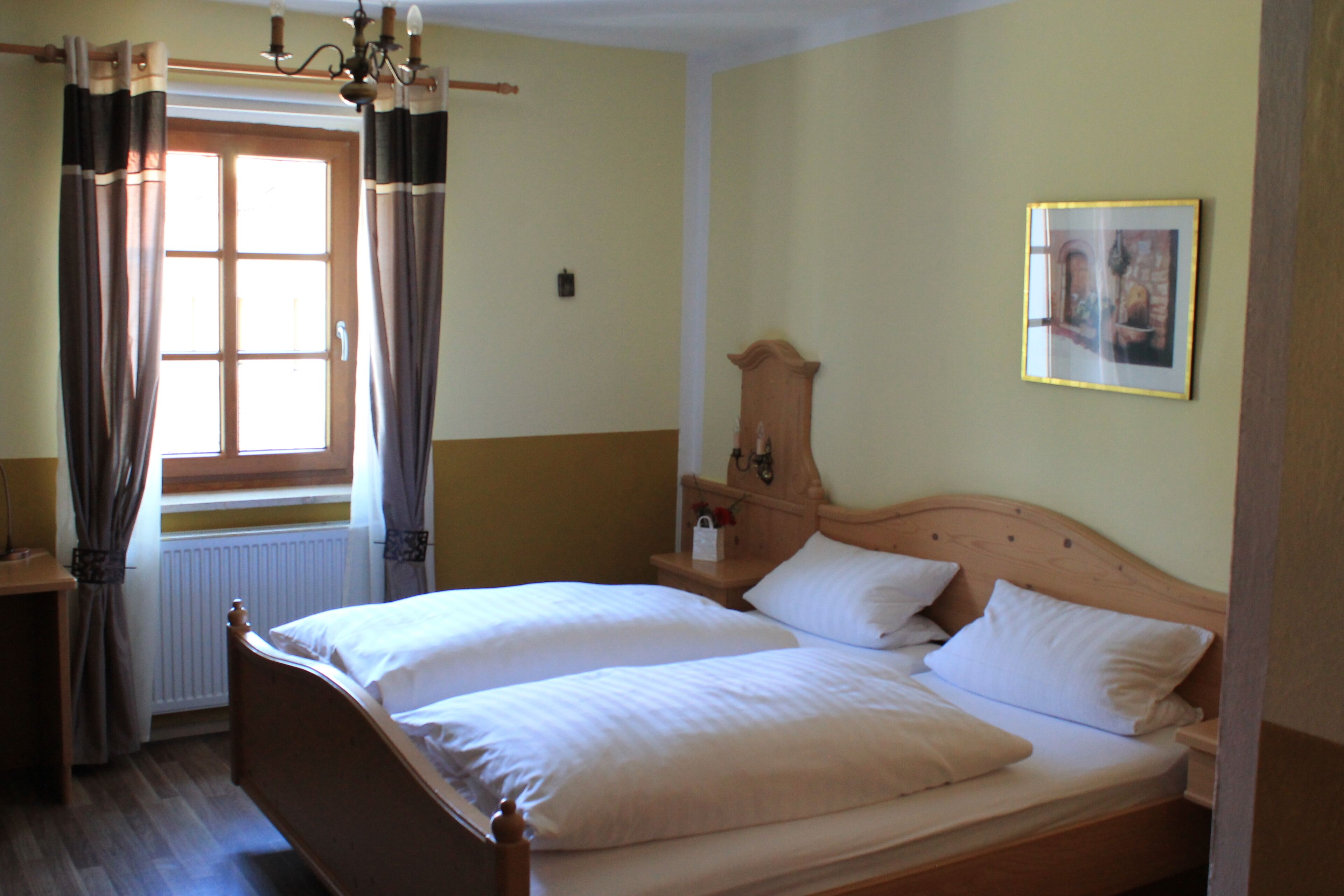A double bed with white linens, a wooden headboard, a nightstand with a lamp, a window with curtains, and a framed picture on the wall.