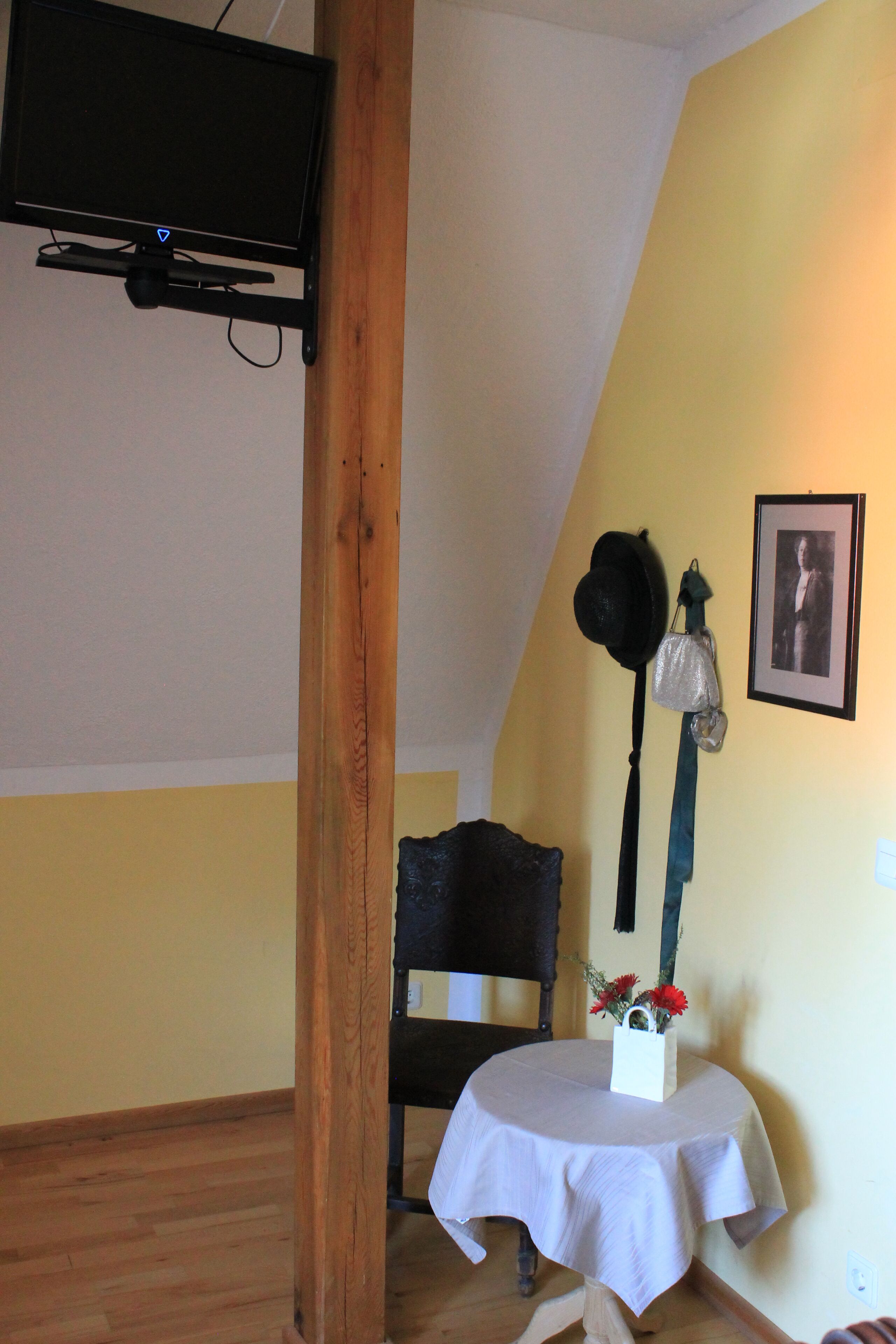 A room with a television mounted on the wall, a wooden chair, a small table with a white tablecloth, and a framed picture on the wall.