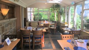 A restaurant interior with tables and chairs, a stone wall, and large windows with a view of greenery. - Calf's Head Hotel (Clitheroe)