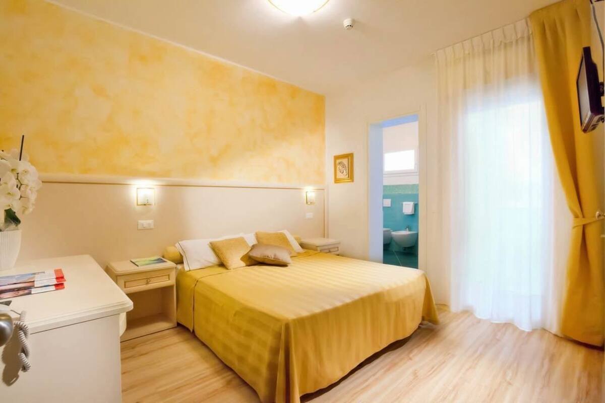Habitación de hotel con cama, mesita de noche, lámpara y vista al baño.