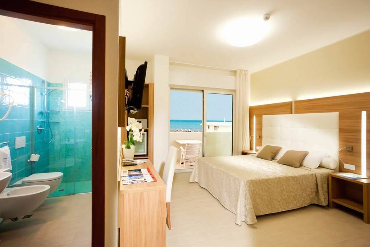 Habitación de hotel con cama, mesita de noche y escritorio con silla. Incluye baño con inodoro y ducha. La habitación tiene vista al mar.