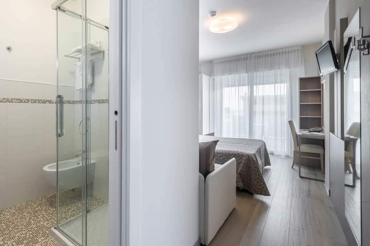 Una habitación de hotel moderna con cabina de ducha de vidrio, una cama con colcha estampada, un escritorio con silla y un televisor montado en la pared.
