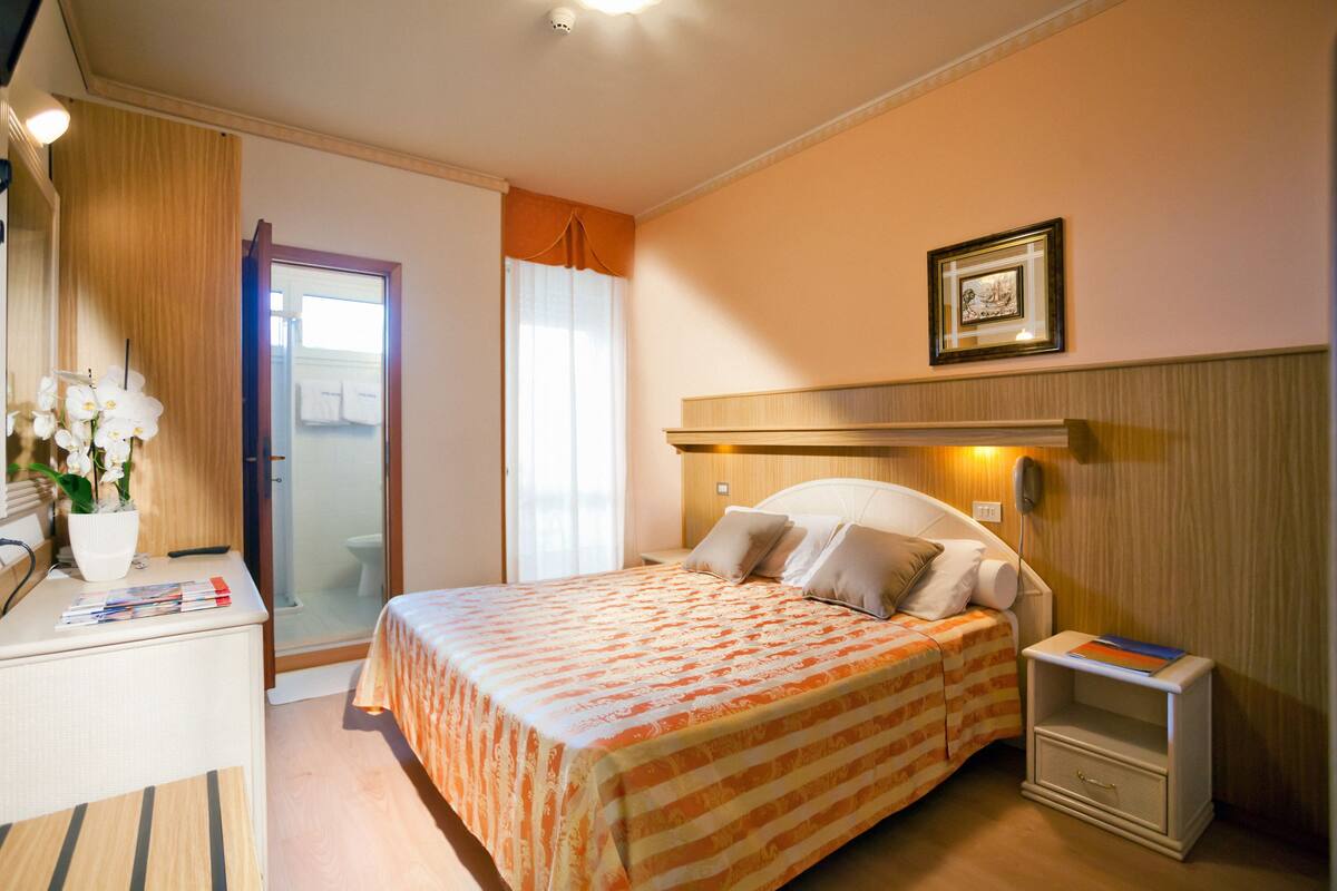 Una habitación de hotel con una cama, mesita de noche y un escritorio pequeño con lámpara y un jarrón de flores.