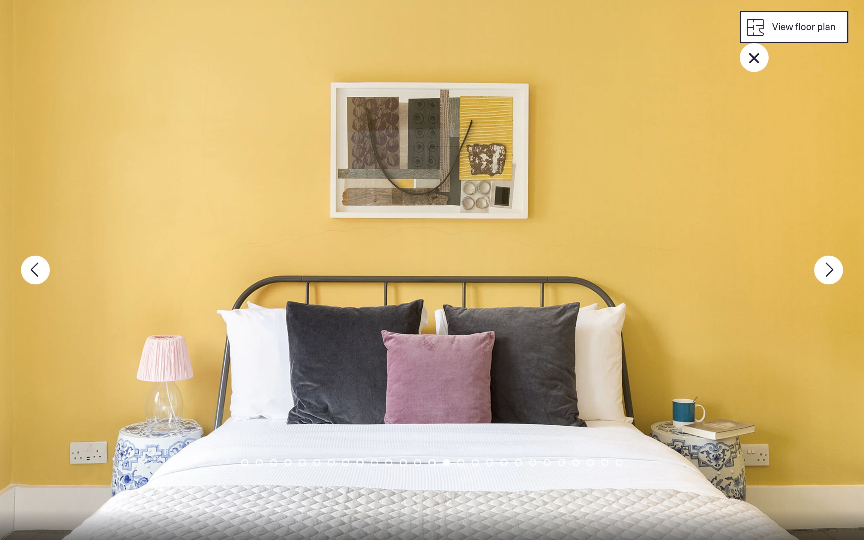 A bed with a white quilt, black and pink pillows, and a framed artwork above it. There are bedside tables with lamps and a cup on one side.