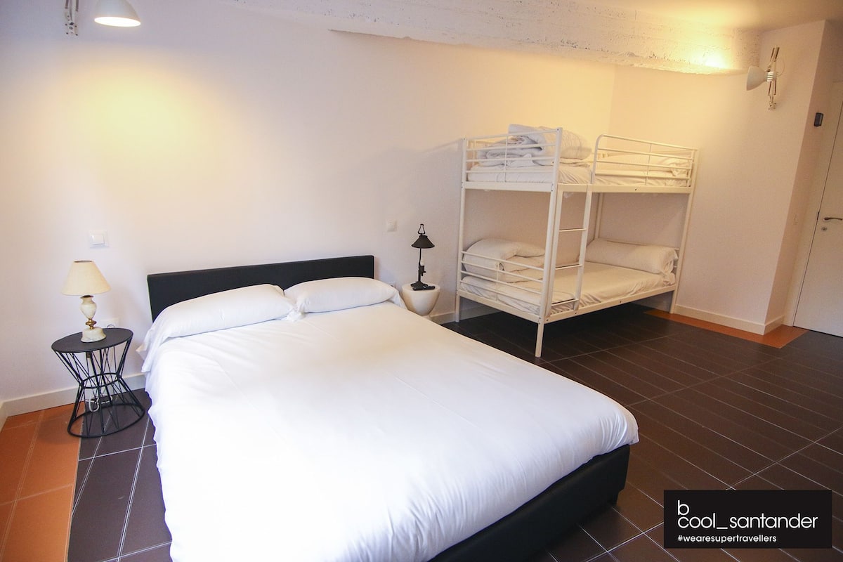 Una cama individual con ropa de cama blanca, una mesita de noche con lámpara, y una cama litera con ropa de cama blanca en una habitación con suelo de baldosas.