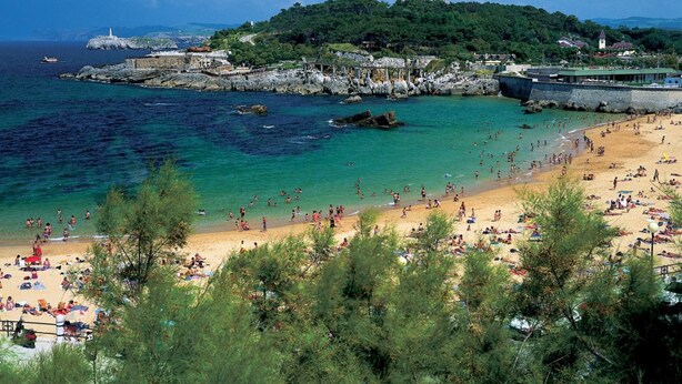 Une plage côtière aux eaux turquoise et limpides, un rivage sablonneux et une colline parsemée de bâtiments, avec un phare en arrière-plan.