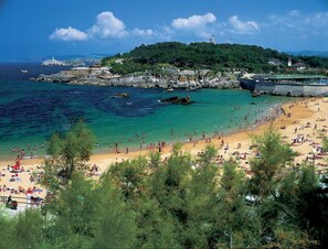 A coastal beach with clear turquoise waters, a sandy shore, and a hill with buildings and a lighthouse in the background. - Enjoy Santander - Hostel (Santander)