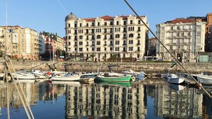 Un port de plaisance avec des bateaux amarrés, un mur de pierre et, au fond, un grand bâtiment doté de nombreuses fenêtres et de balcons.