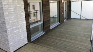 A covered outdoor area with wooden decking, a brick wall, and large sliding glass doors. - Fornebu Apartments - Fornebuporten (Bærum)