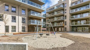 A multi-story residential building with balconies and a playground area with swings and a slide. - Fornebu Apartments - Fornebuporten (Bærum)