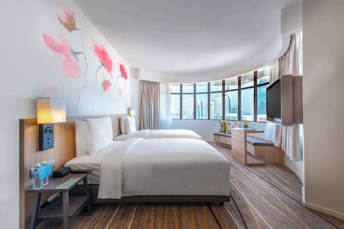 A hotel room with two beds, a desk, a TV, and a large window with a city view.