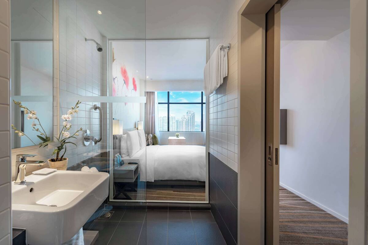 A modern hotel bathroom with a glass shower enclosure, a large mirror, a sink, and a bedside table with a plant.