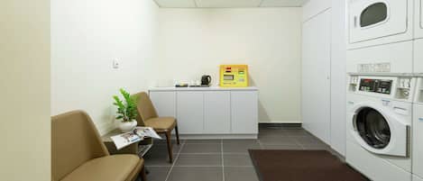 A small laundry room with a washing machine, a dryer, and a control panel. There are two chairs, a small table with a plant, and a magazine.
