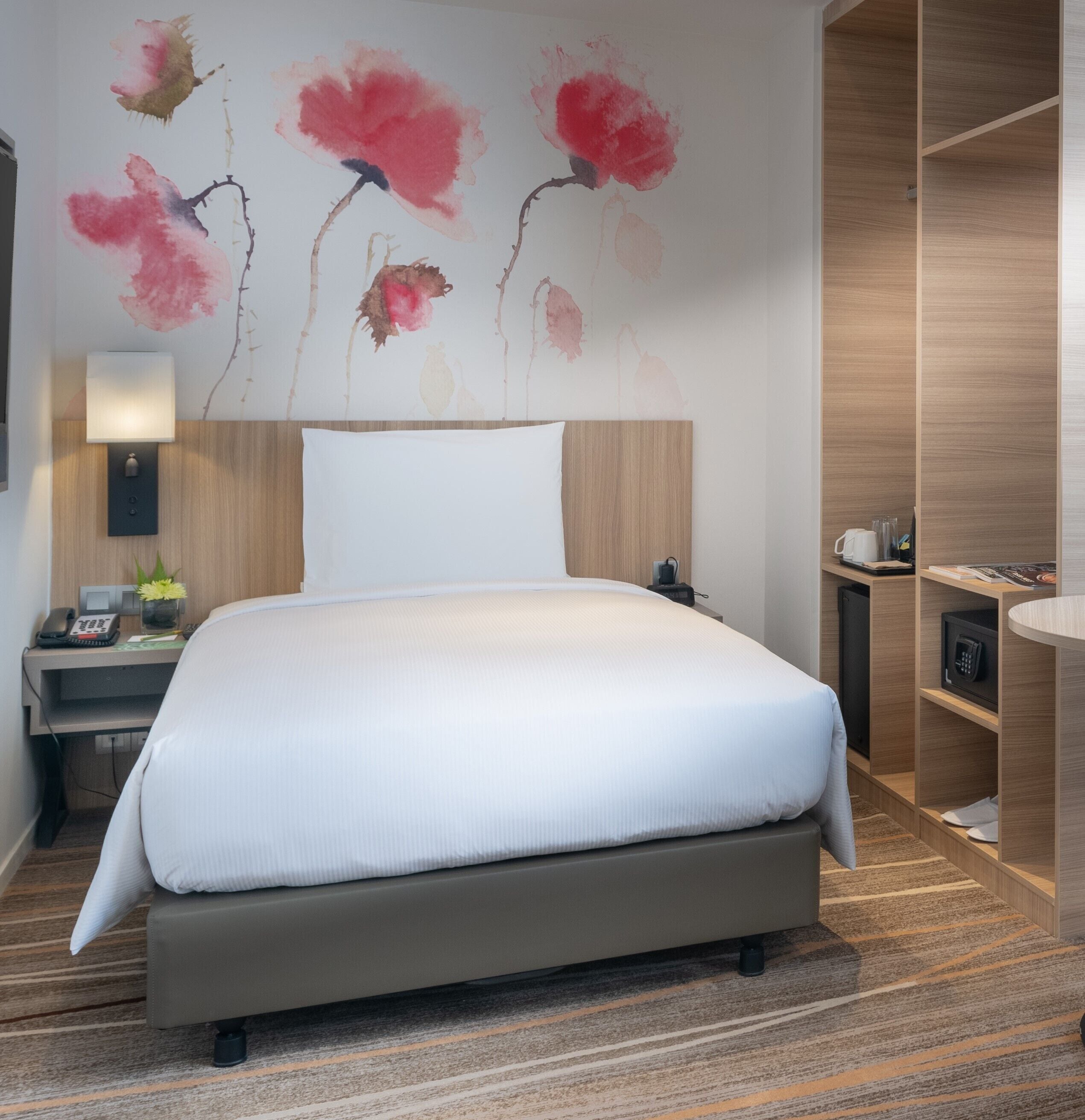 A hotel room with a large bed, a bedside table with a lamp, a phone, and a small plant. There is a wooden wardrobe and a desk with a chair.
