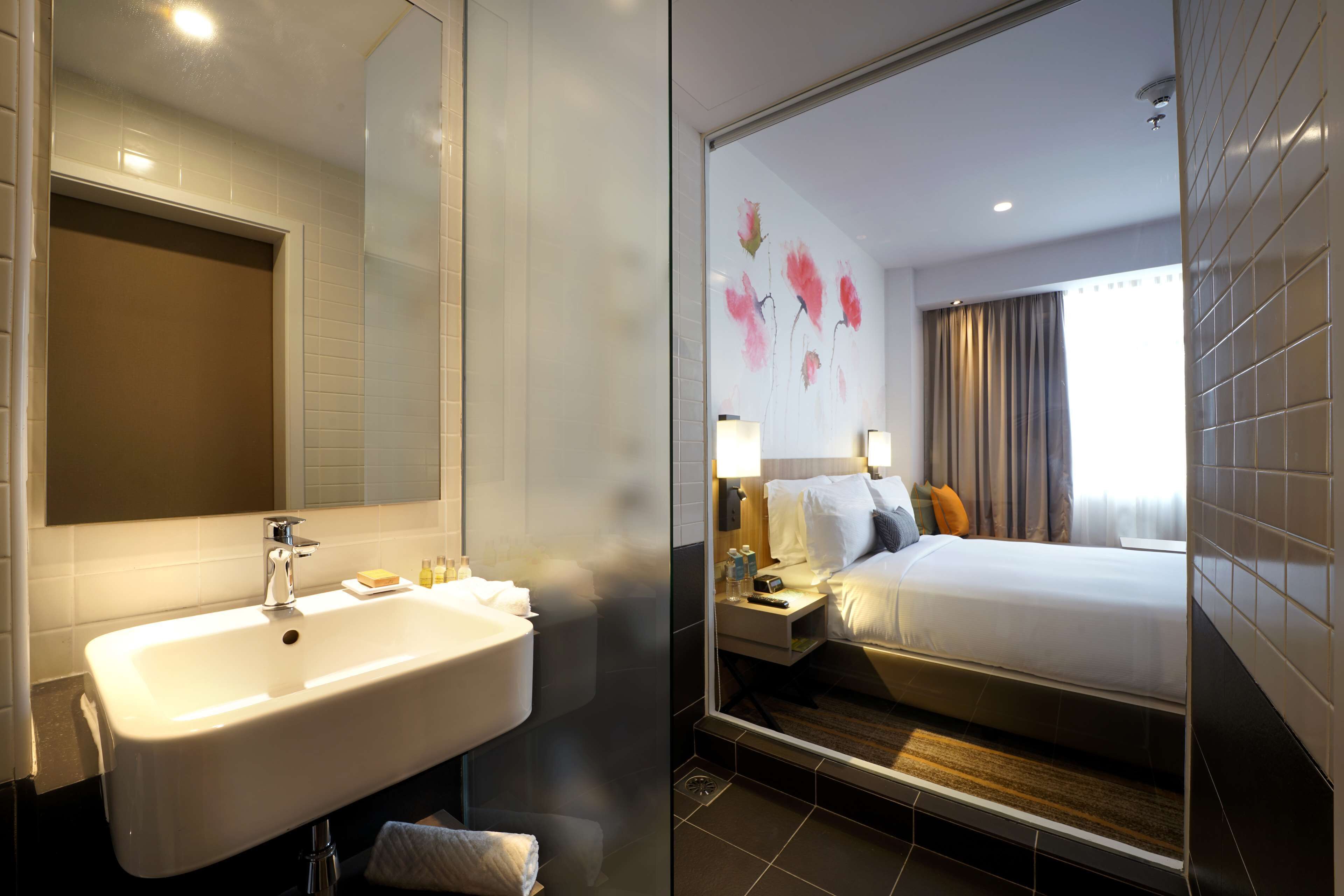 A hotel bathroom with a sink, mirror, and a shower. The room has a bed with pillows and a nightstand with a lamp.