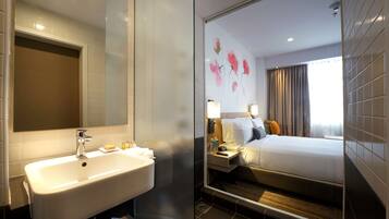 A hotel bathroom with a sink, mirror, and a shower. The room has a bed with pillows and a nightstand with a lamp.