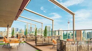 A rooftop terrace with wicker furniture, a view of the city skyline, and a modern architectural structure. - Hilton Garden Inn Kuala Lumpur Jalan Tuanku Abdul Rahman South (Kuala Lumpur)