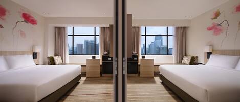 A hotel room with two beds, a desk, and a view of the cityscape through large windows.