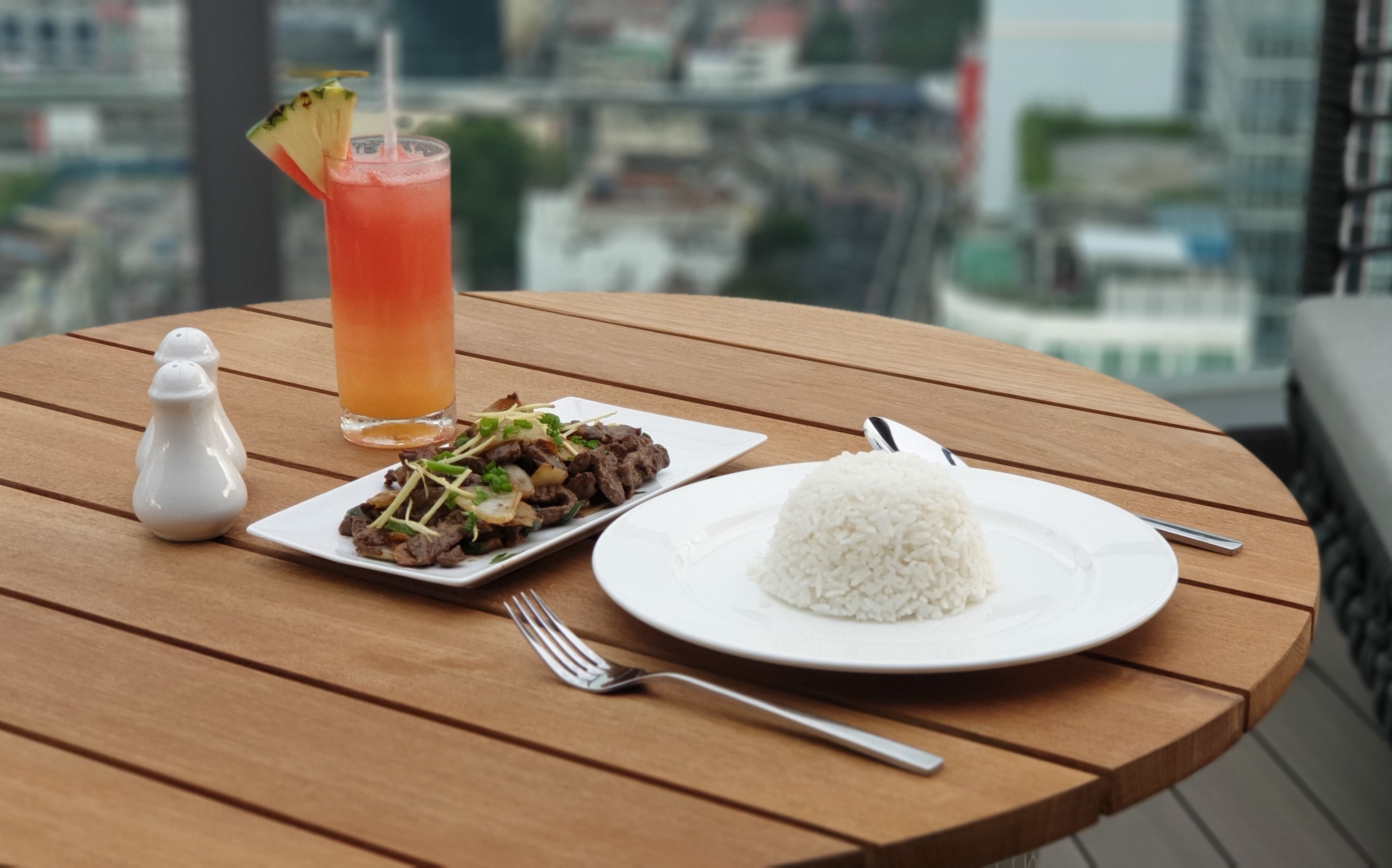 Uma mesa com uma refeição de carne salteada, arroz branco e um copo de bebida vermelha com enfeite.