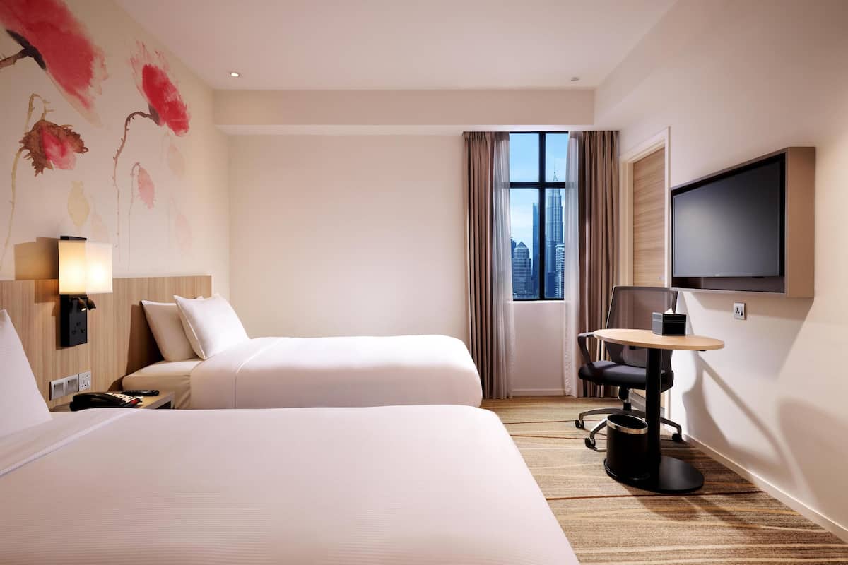 A hotel room with two beds, a desk with a chair, a TV, and a window with a city view.