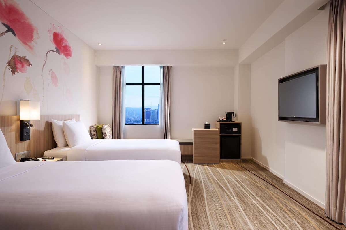 A hotel room with two beds, a desk, a TV, and a large window with a city view.