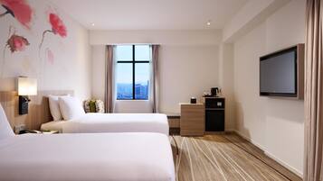 Habitación de hotel con dos camas, un escritorio, un televisor y un amplio ventanal con vistas a la ciudad.