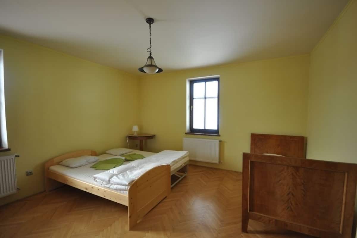 Een kleine kamer met een enkel bed, een houten bed, een klein nachtkastje, een stoel, een raam en een radiator.