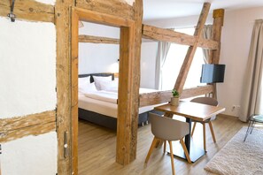 Une pièce avec des poutres apparentes, un lit, un bureau avec une chaise, une télévision et une petite plante.