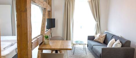 A modern living room with a wooden accent wall, a grey sofa, a wooden dining table with a chair, a TV mounted on the wall, and a large window with curtains.