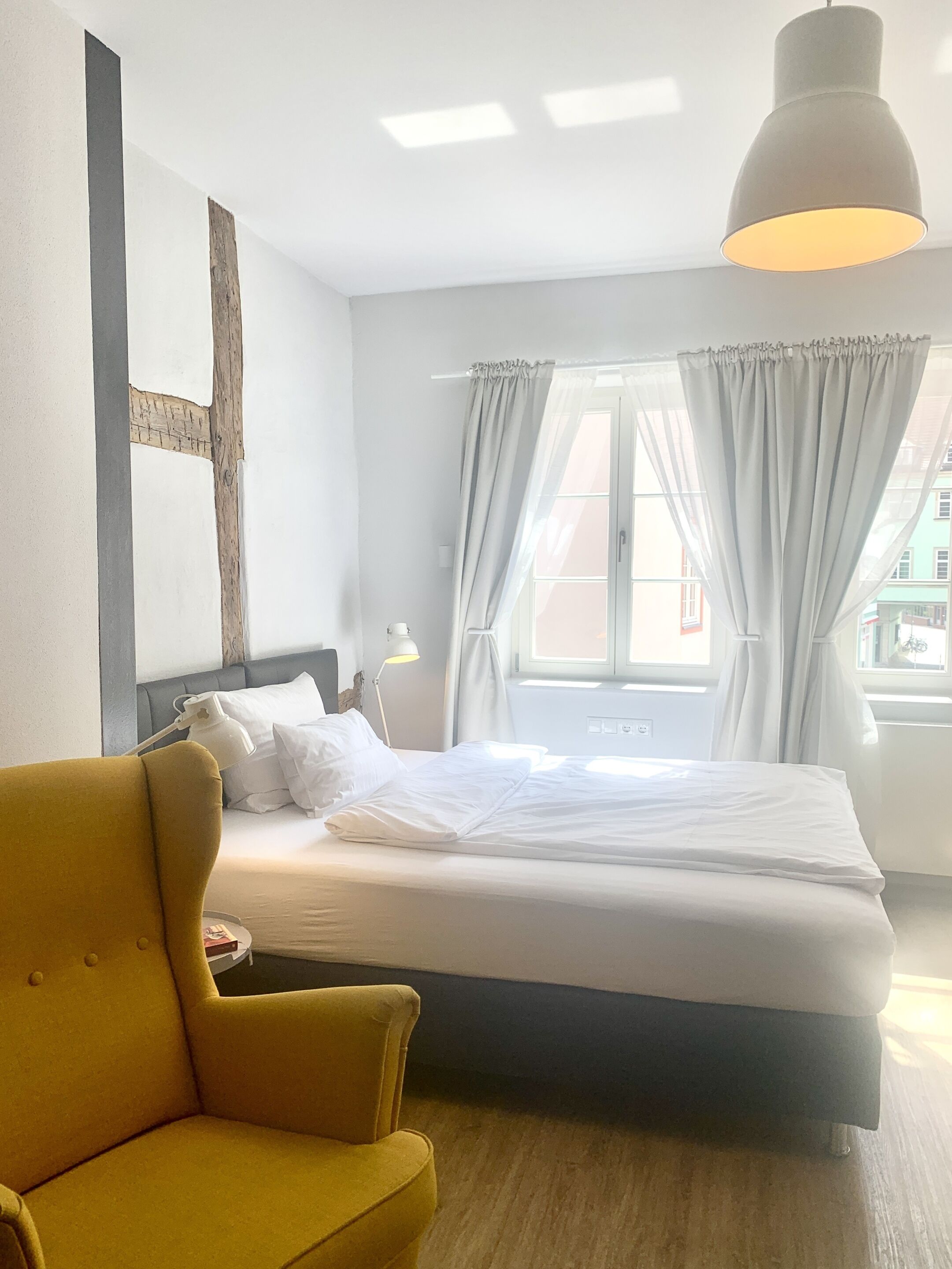 A hotel room with a bed, a yellow armchair, a bedside table with a lamp, and a large window with curtains.