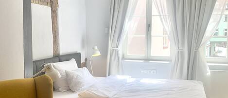A hotel room with a bed, a yellow armchair, a bedside table with a lamp, and a large window with curtains.