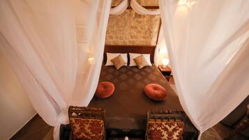 Quarto com cama de dossel, duas almofadas, mesa de cabeceira com garrafa e copos, e duas poltronas estofadas.