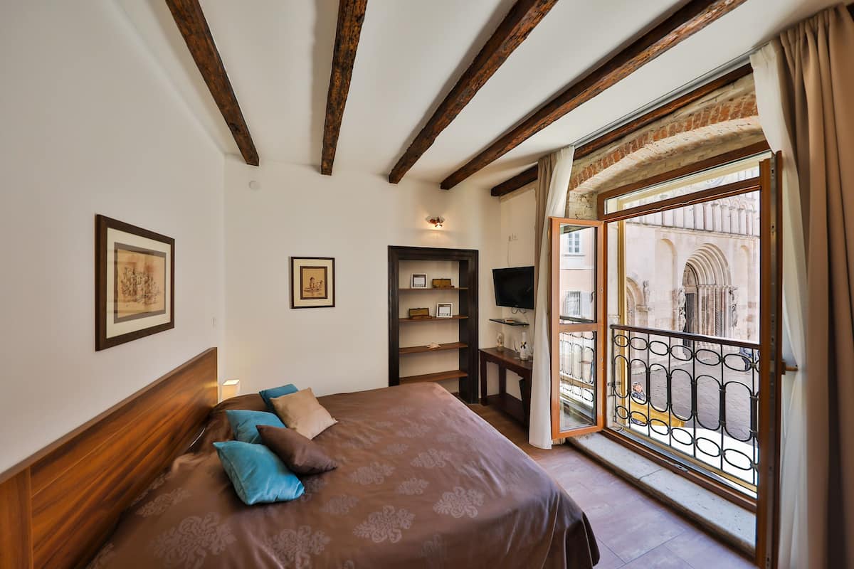 Un dormitorio con una cama grande, cabecera de madera, televisor, estantería y balcón con barandilla de hierro forjado.