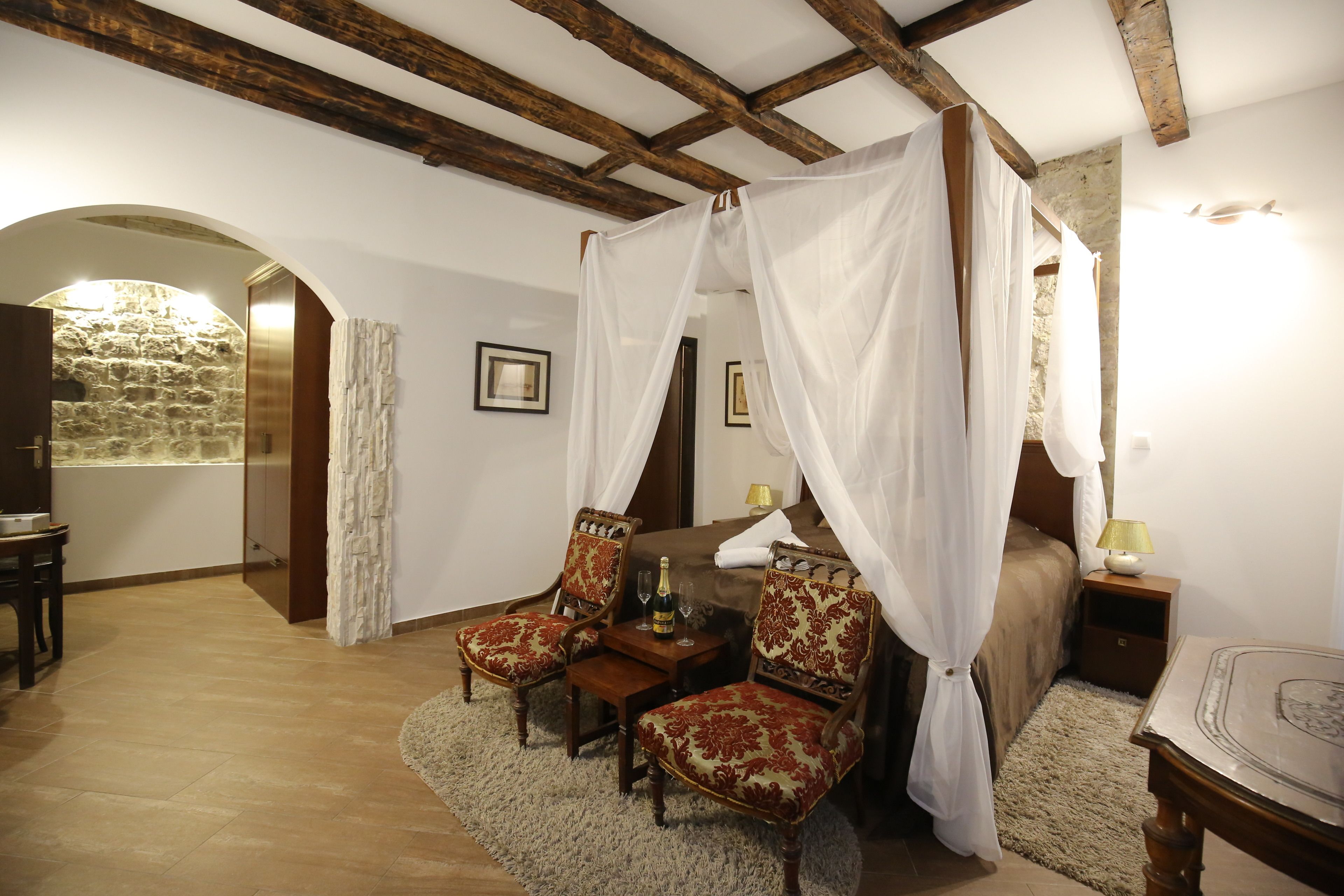 Una camera da letto con un letto a baldacchino, due poltrone imbottite, un tavolino con una bottiglia e un comodino con una lampada.