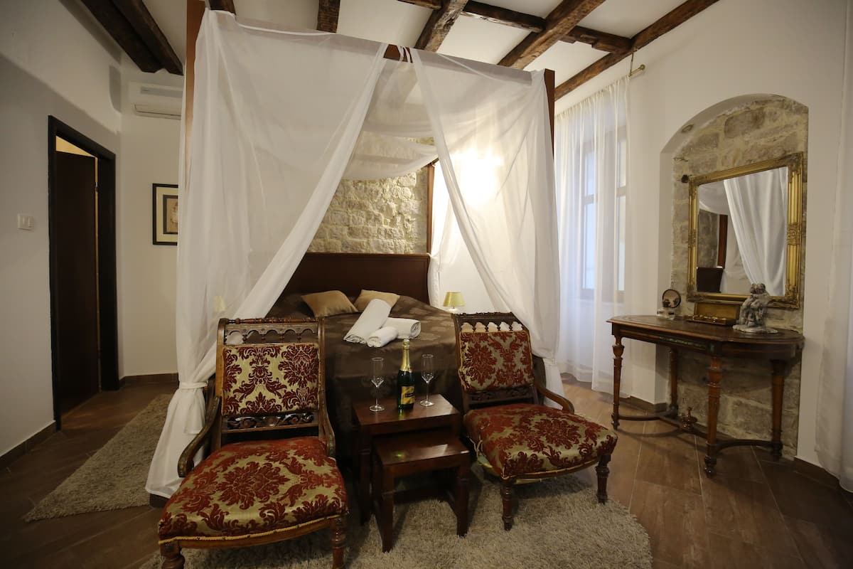 Un dormitorio con una cama con dosel, una mesita de noche, dos sillones tapizados, una mesa de madera y un espejo.