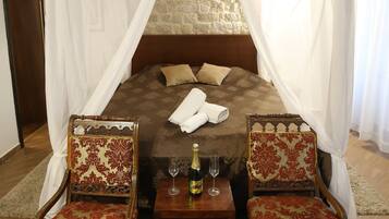 Une chambre à coucher avec un lit à baldaquin, un dais, deux fauteuils rembourrés, une petite table avec une bouteille et un verre, et un mur de pierre derrière le lit.