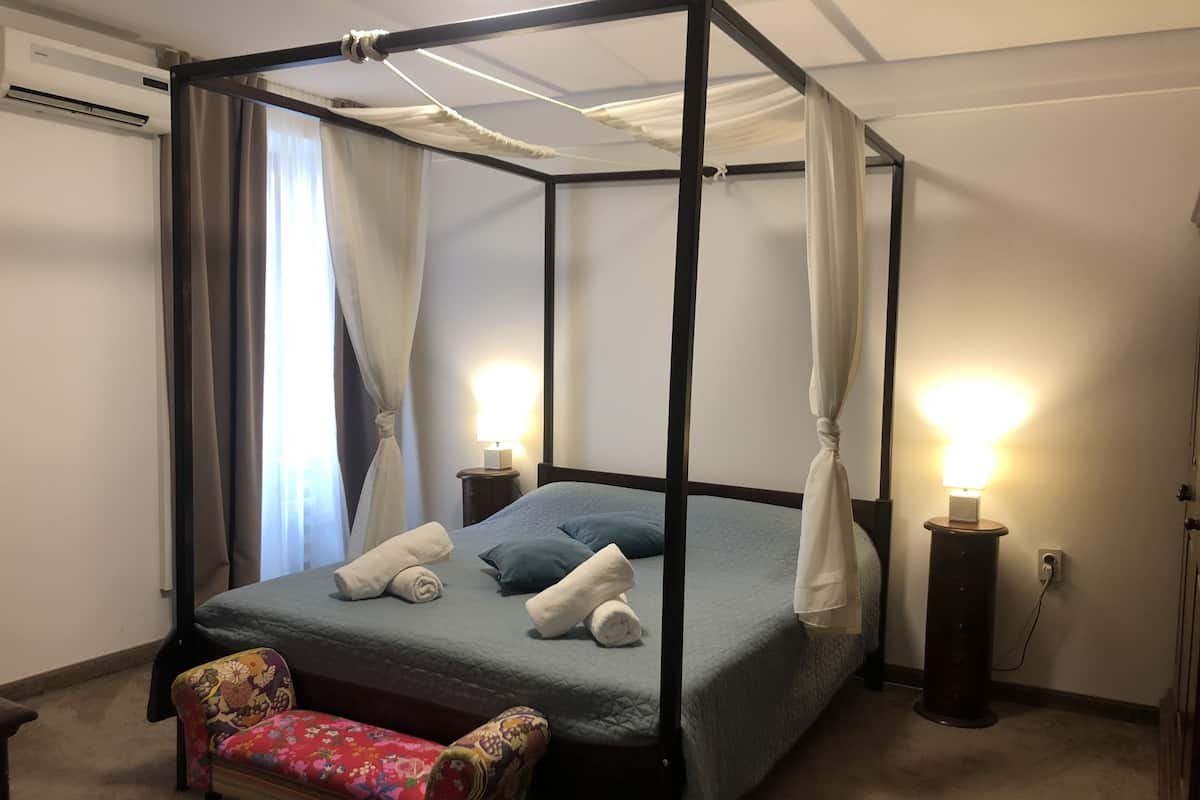 Una cama con dosel, dos lámparas de noche, un banco floral y una mesita con lámpara.