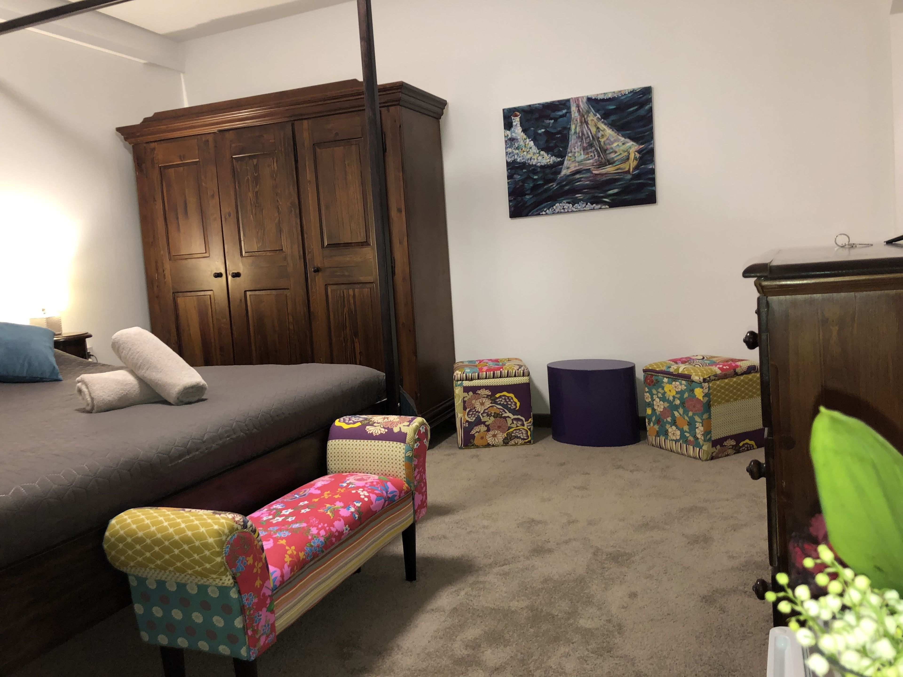 Une chambre à coucher avec un lit, une armoire en bois, un banc, deux petits tabourets et un tableau accroché au mur.