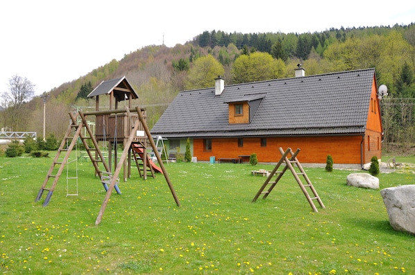 A wooden play structure and a house with a sloped roof in a grassy field.