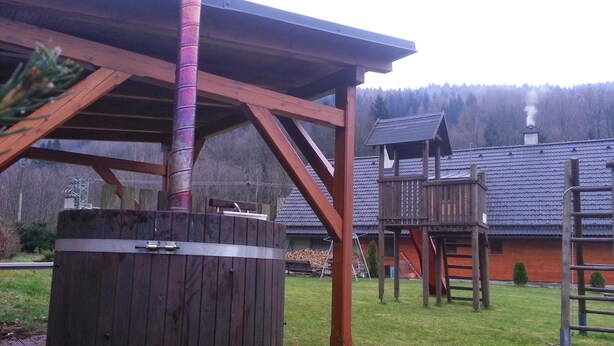 A wooden hot tub with a chimney, a wooden deck, and a play structure in a backyard. - Chata Pod Vlekem (Loucna nad Desnou)