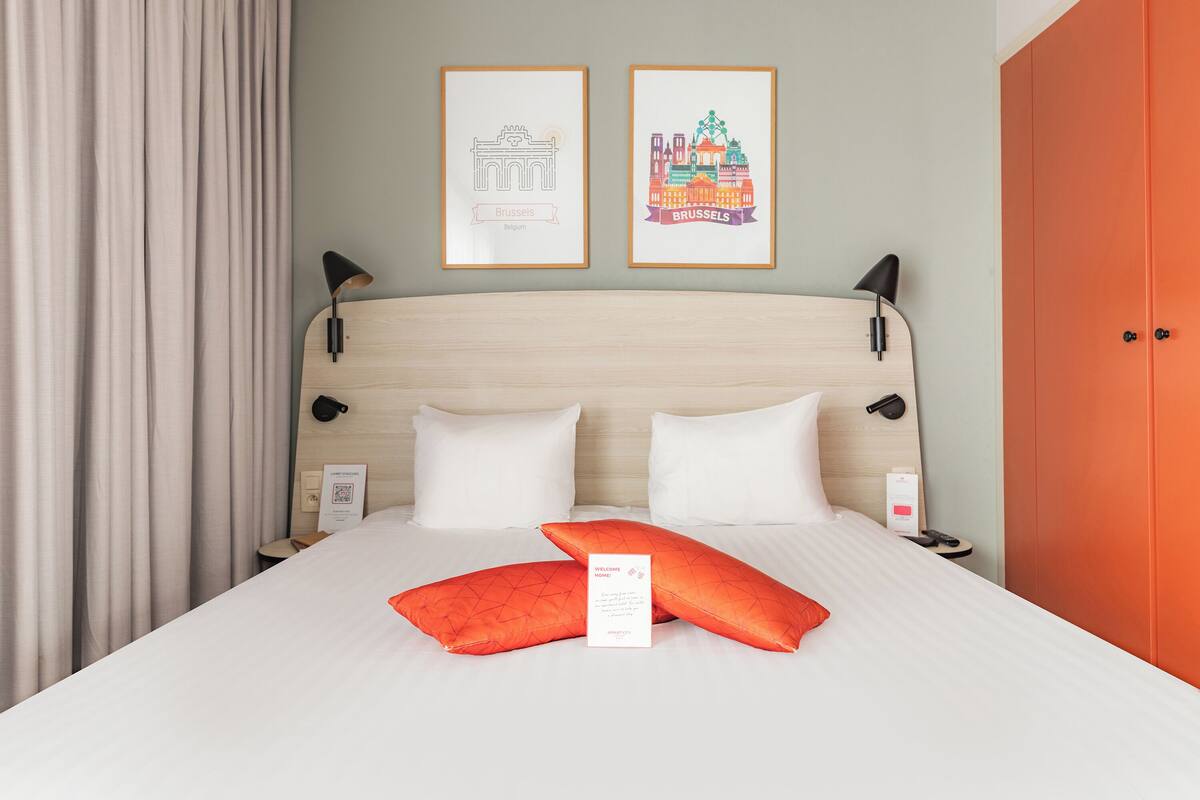 Ein Hotelzimmer mit Bett, Kissen, einem gerahmten Willkommensschild, einem Nachttisch, einer Lampe und einer Tür.
