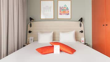 A hotel room with a bed, pillows, a framed welcome sign, a nightstand, a lamp, and a door.