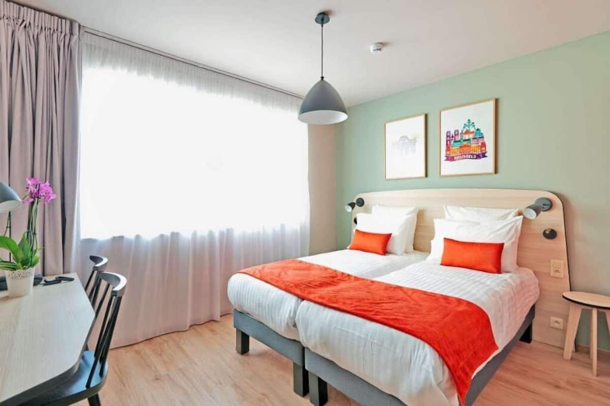 Ein Doppelbett mit weißer und orangefarbener Bettwäsche, ein Esstisch mit Stühlen, eine hängende Pendelleuchte und zwei gerahmte Bilder an der Wand.