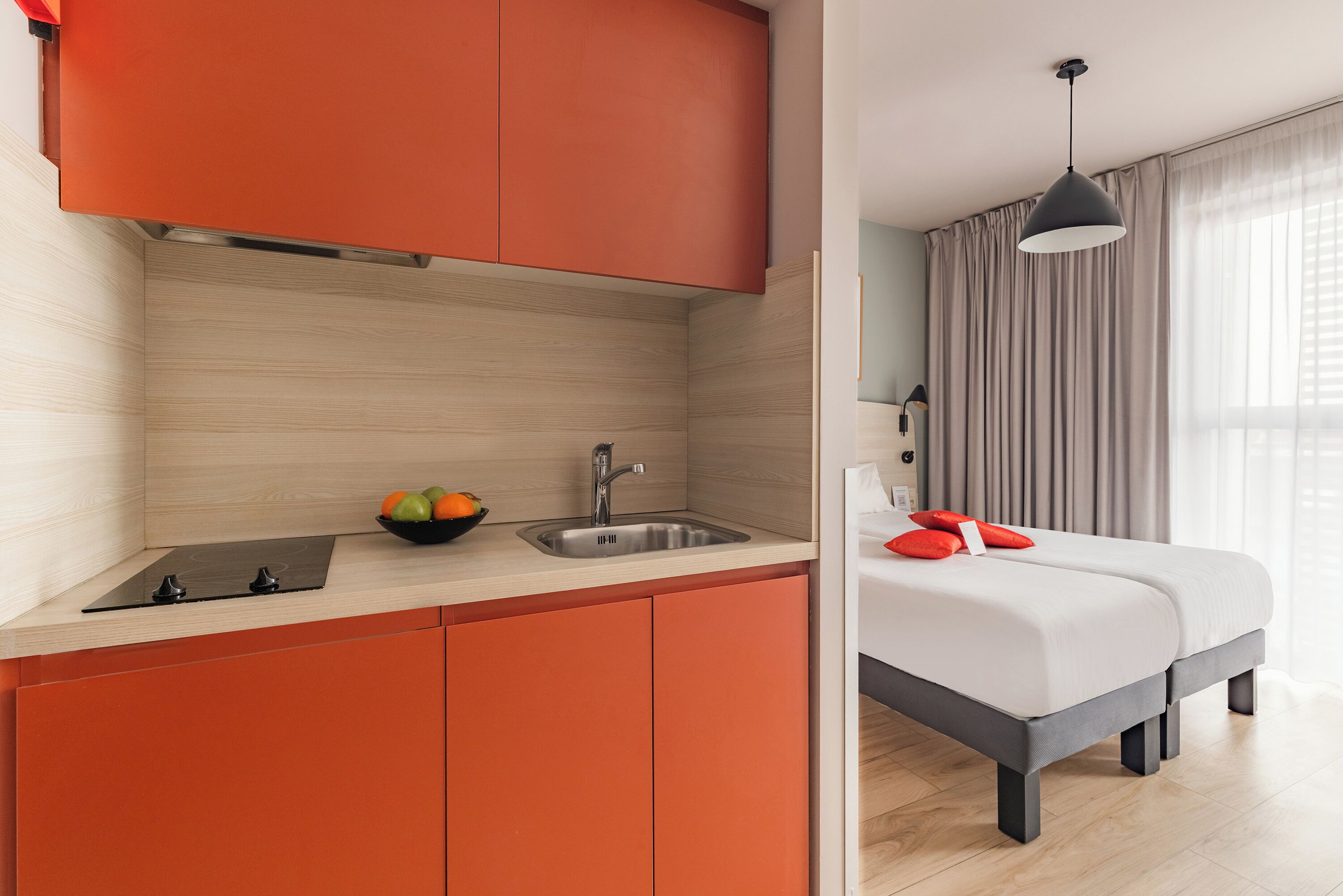 Une pièce compacte avec une kitchenette, un lit agrémenté d’oreillers rouges et une fenêtre avec des rideaux.
