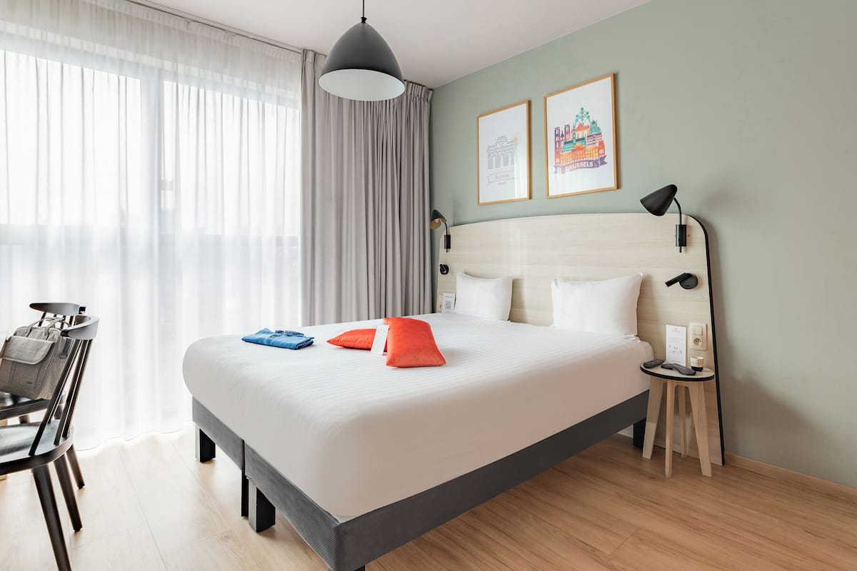 Ein Hotelzimmer mit einem Bett, einem Schreibtisch, einem Sessel sowie einer Lampe und zwei gerahmten Bildern an der Wand.