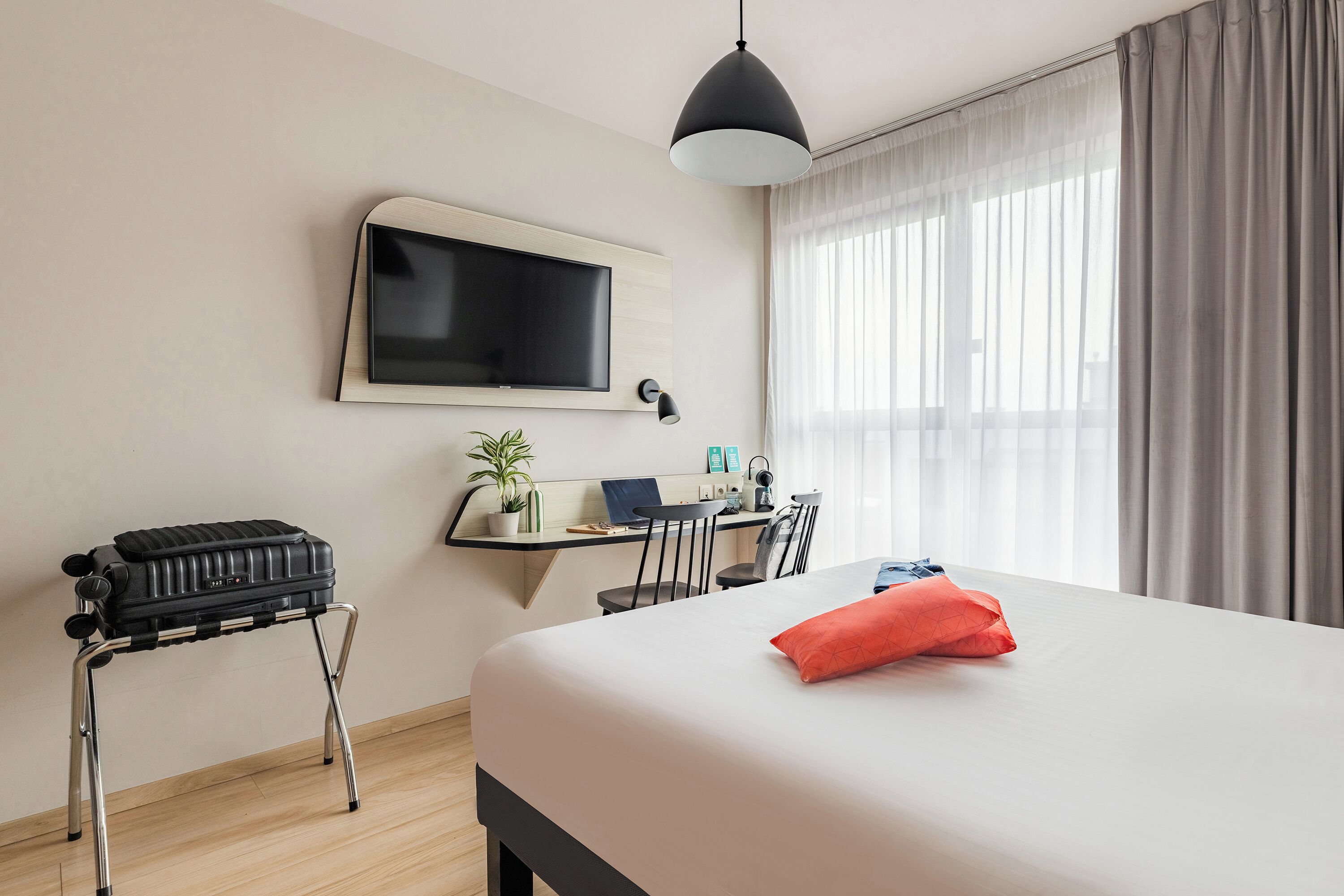 A hotel room with a bed, a desk with a computer, a television, and a suitcase on a stool.