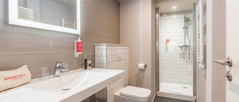 Un baño moderno con un lavabo blanco, espejo e inodoro, una zona de ducha con puerta de cristal y un perchero para toallas.