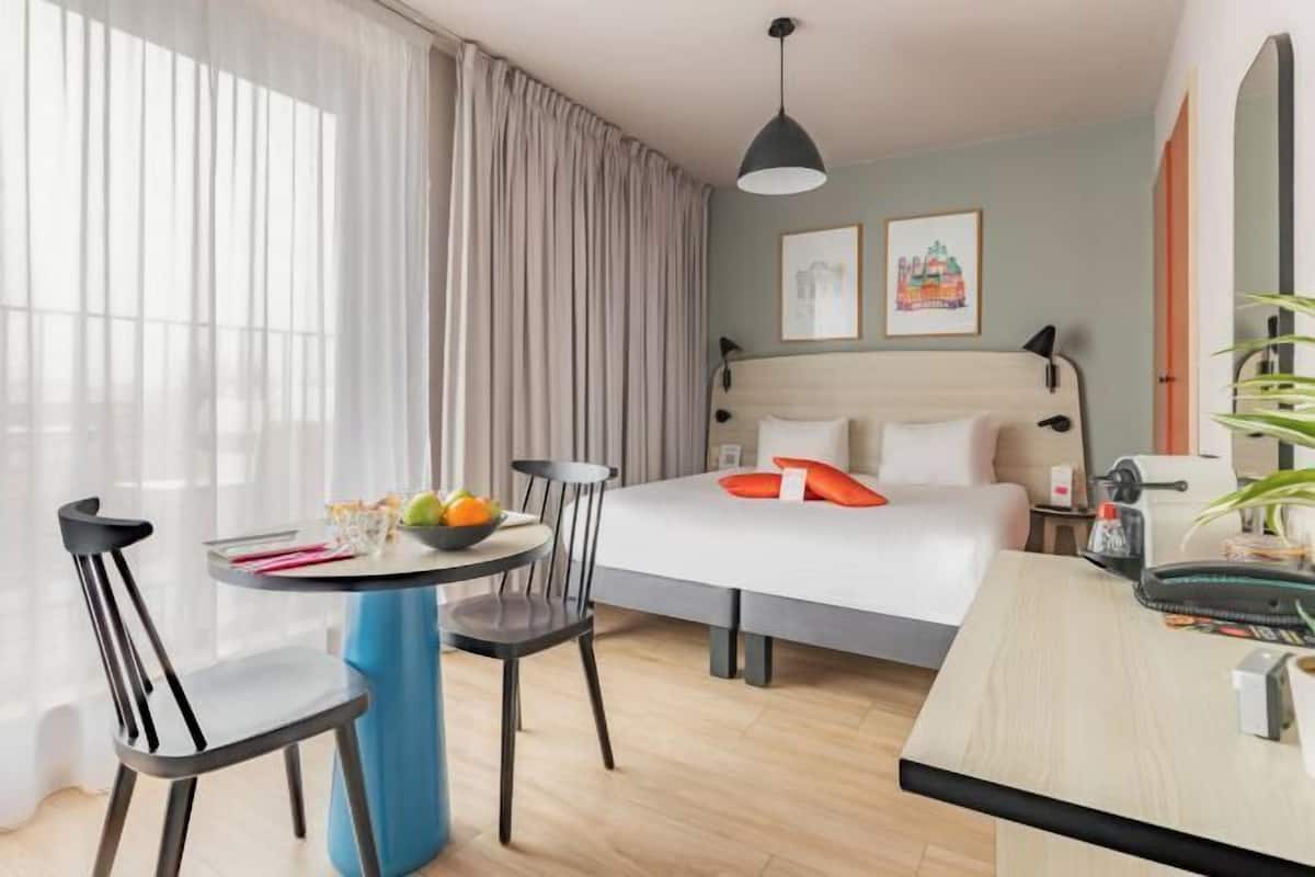 Ein modernes Hotelzimmer mit einem Bett, einem Esstisch, Stühlen, einer kleinen Küchenzeile und einem großen Fenster mit Vorhängen.