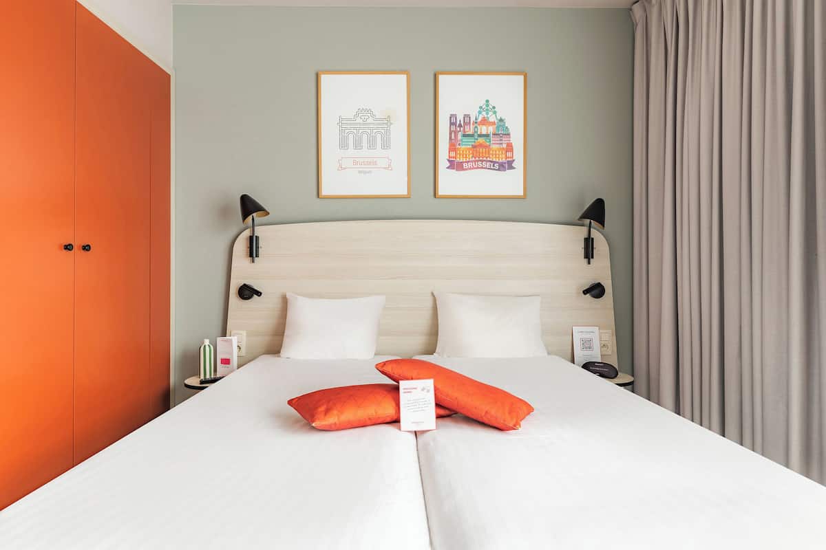 Ein Hotelzimmer mit einem Bett, zwei gerahmten Bildern, einem Nachttisch, einer Lampe und einem Vorhang.