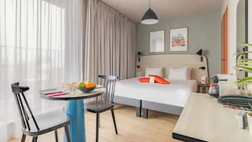 Una habitación de hotel moderna con cama, escritorio, silla, mesa con fruta y un ventanal con cortinas.